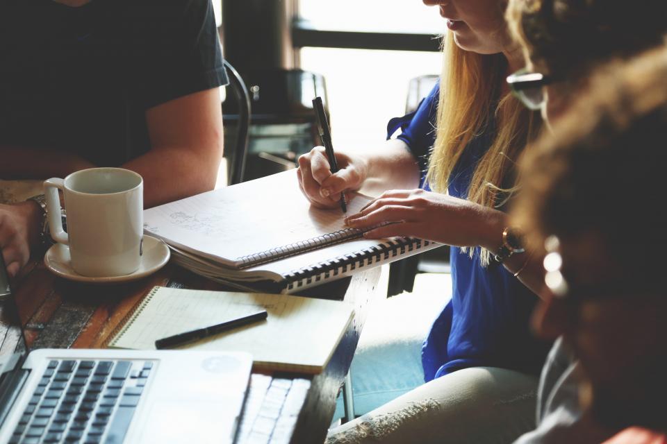 People at a table, one writing notes in a spiral notebook next to a cup of coffee