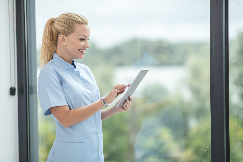 A nurse in a blue uniform reading a tablet by a window