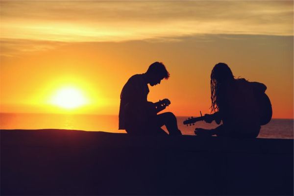 Duas pessoas tocam violão numa praia ao pôr do sol, em contraluz
