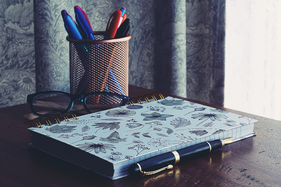 Un cuaderno cerrado con una pluma, unas gafas y un lapicero sobre un escritorio