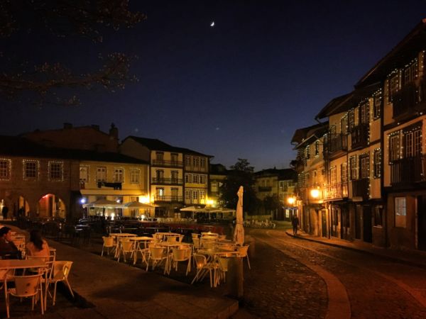 Una plaza de un casco antiguo de noche, con mesas de terraza, casas iluminadas y la luna