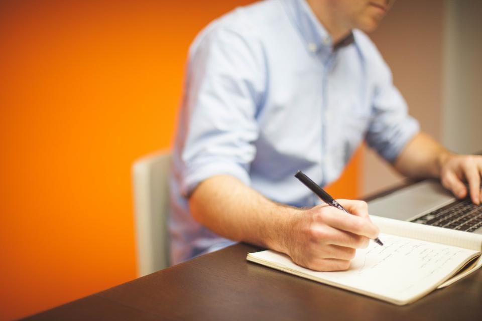 A person writing by hand at a desk next to a laptop
